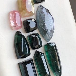 K&ouml;nnte beinhalten: Eine Sammlung facettierter Edelsteine in verschiedenen Formen und Gr&ouml;&szlig;en, darunter Schattierungen in Dunkelgr&uuml;n, Schwarz, Pink, Gelb und ein gro&szlig;er, l&auml;nglicher graublauer Stein. Jeder Stein weist mehrere polierte Oberfl&auml;chen auf, die das Licht reflektieren.