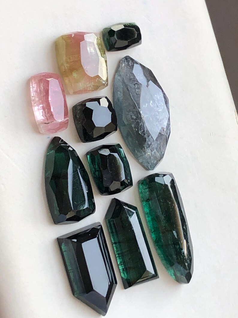 K&ouml;nnte beinhalten: Eine Sammlung facettierter Edelsteine in verschiedenen Formen und Gr&ouml;&szlig;en, darunter Schattierungen in Dunkelgr&uuml;n, Schwarz, Pink, Gelb und ein gro&szlig;er, l&auml;nglicher graublauer Stein. Jeder Stein weist mehrere polierte Oberfl&auml;chen auf, die das Licht reflektieren.