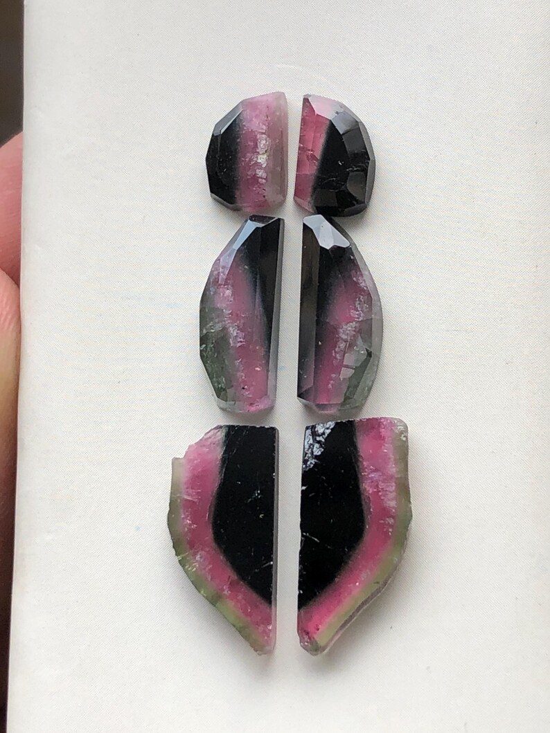 May include: Set of six gemstone cabochons, featuring a mix of black, pink, and green colors. The cabochons are cut in a variety of shapes, including ovals and half-ovals.