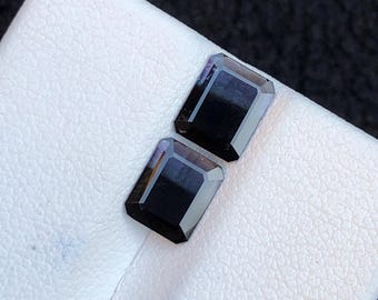 May include: Two rectangular, dark gemstones with a polished, reflective surface. The gems are set on a white, textured surface, with a dark background. The gems have a deep, black color and an emerald cut.