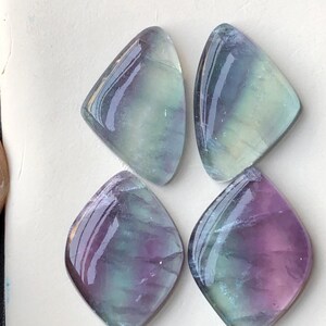 May include: Four polished fluorite gemstones. Each stone has a unique shape, with a rounded top and a pointed bottom. The stones display a mix of colours, including purple, green, and blue, with a glossy finish. The gemstones are arranged on a white surface.