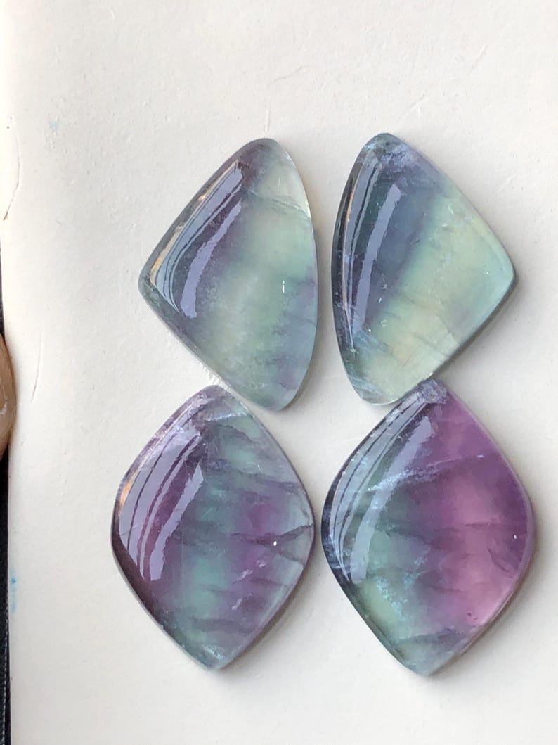 May include: Four polished fluorite gemstones. Each stone has a unique shape, with a rounded top and a pointed bottom. The stones display a mix of colours, including purple, green, and blue, with a glossy finish. The gemstones are arranged on a white surface.
