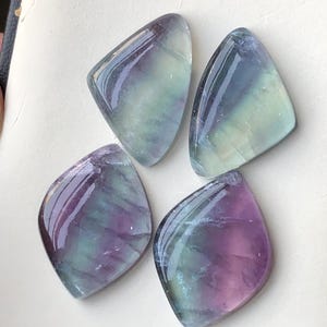 May include: Four polished fluorite gemstones in shades of purple, green, and blue. Each stone has a rounded, triangular shape with a glossy surface. The gemstones are arranged on a white surface.