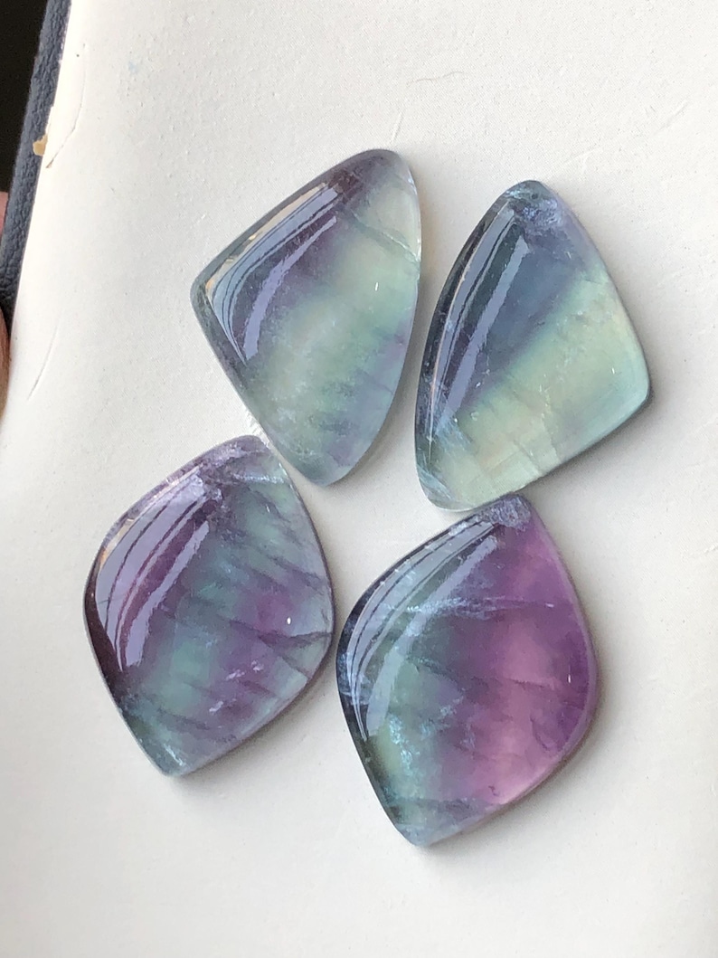 May include: Four polished fluorite gemstones in shades of purple, green, and blue. Each stone has a rounded, triangular shape with a glossy surface. The gemstones are arranged on a white surface.