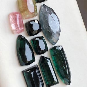 K&ouml;nnte beinhalten: Eine Sammlung von zehn facettierten Edelsteinen in verschiedenen Formen und Gr&ouml;&szlig;en. Die Steine zeigen Schattierungen von tiefem Waldgr&uuml;n, durchscheinendem Rosa, blassem Gelbgr&uuml;n und dunklem Anthrazit, alle mit polierten, geometrisch facettierten Oberfl&auml;chen.