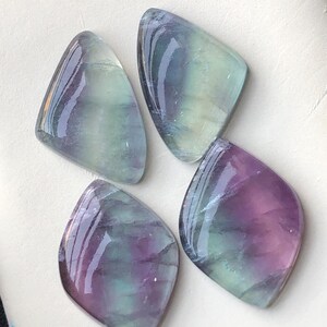 May include: Four polished fluorite gemstones in shades of purple, green, and clear. Each stone has a unique shape, with smooth, reflective surfaces. The gemstones are arranged on a white surface, showcasing their natural beauty.