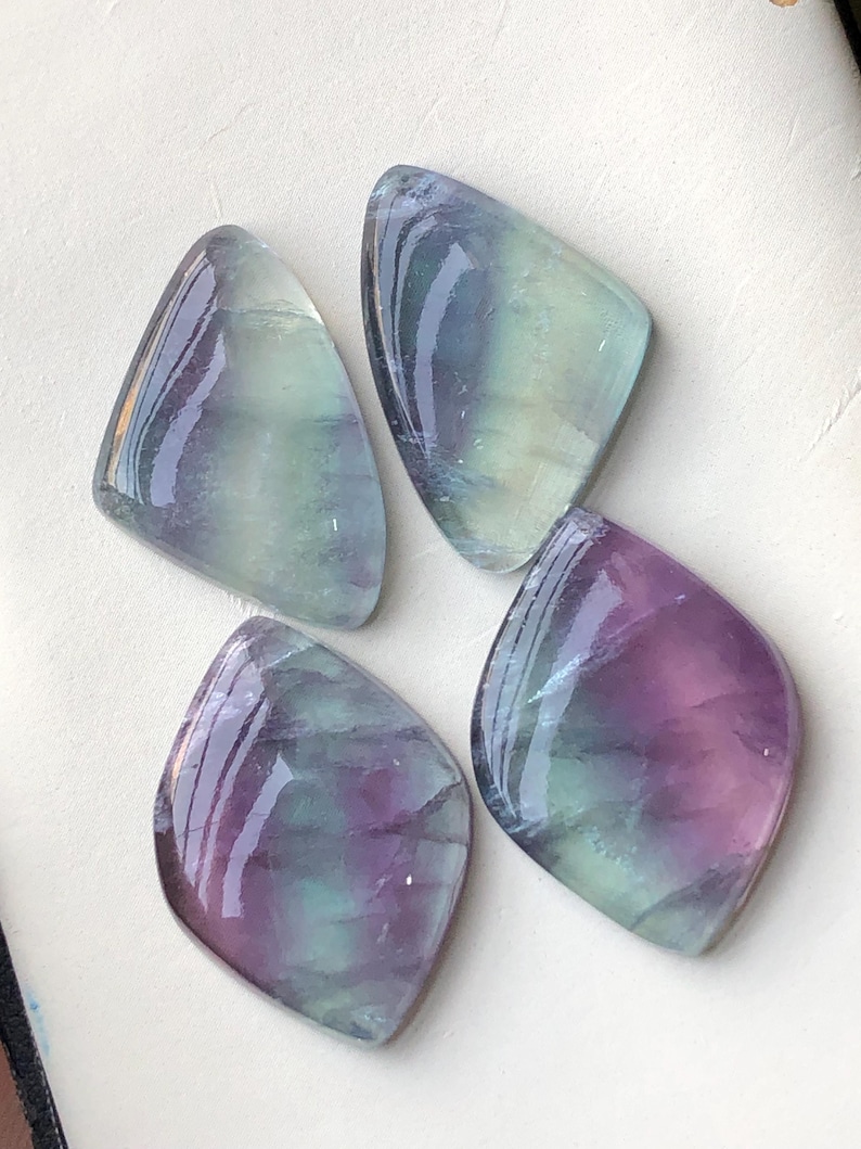 May include: Four polished fluorite gemstones in shades of purple, green, and clear. Each stone has a unique shape, with smooth, reflective surfaces. The gemstones are arranged on a white surface, showcasing their natural beauty.