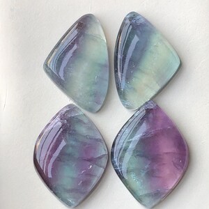 May include: Four polished fluorite gemstones. Each stone is a unique shape with a rounded bottom and a pointed top. The stones display a mix of purple, green, and blue hues, with a glossy finish. The gemstones are arranged on a white surface.