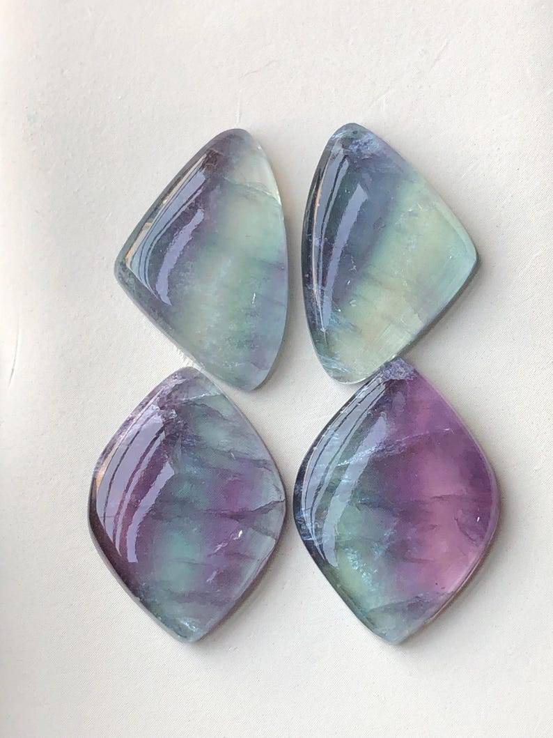 May include: Four polished fluorite gemstones. Each stone is a unique shape with a rounded bottom and a pointed top. The stones display a mix of purple, green, and blue hues, with a glossy finish. The gemstones are arranged on a white surface.