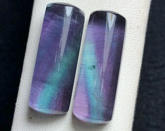 May include: Two rectangular polished fluorite cabochons featuring vibrant bands of deep purple and bright teal. The stones have a smooth, glossy surface with visible internal color zoning and natural mineral inclusions throughout the translucent material.