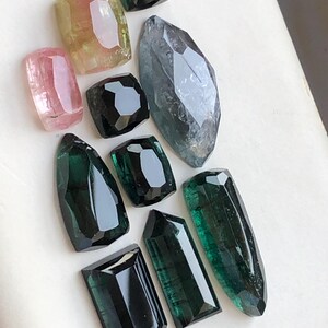 K&ouml;nnte beinhalten: Eine Sammlung von zehn facettierten Edelsteinen in verschiedenen Formen und Gr&ouml;&szlig;en, darunter Schattierungen in Rosa, Gelb, Dunkelgr&uuml;n und Schwarz. Die Steine haben polierte Oberfl&auml;chen mit geometrischen Schliffen und zeigen durchscheinende T&ouml;ne.