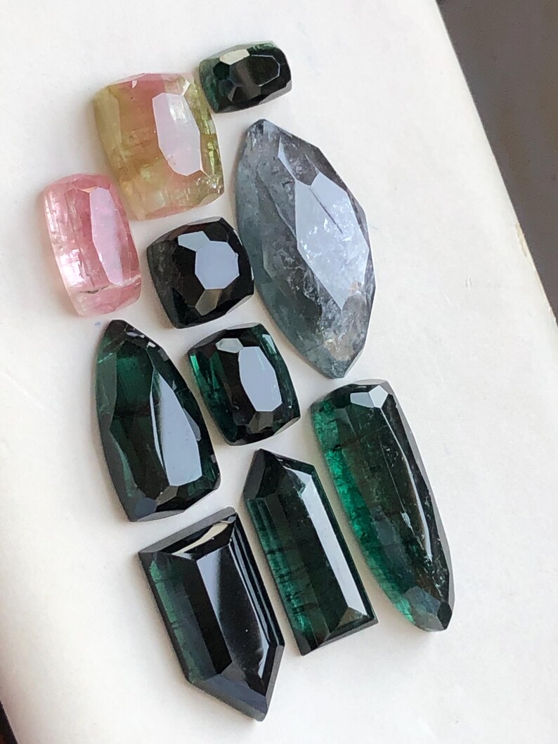 K&ouml;nnte beinhalten: Eine Sammlung von zehn facettierten Edelsteinen in verschiedenen Formen und Gr&ouml;&szlig;en, darunter Schattierungen in Rosa, Gelb, Dunkelgr&uuml;n und Schwarz. Die Steine haben polierte Oberfl&auml;chen mit geometrischen Schliffen und zeigen durchscheinende T&ouml;ne.