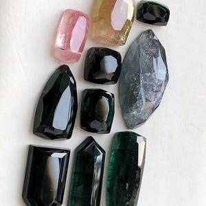 K&ouml;nnte beinhalten: Eine Sammlung von zehn facettierten Edelsteinen in verschiedenen Formen und Gr&ouml;&szlig;en. Die Steine zeigen Schattierungen von tiefem Schwarz, dunklem Waldgr&uuml;n, durchscheinendem Rosa und einem blassen Gelb-Rosa. Jeder Stein ist poliert.