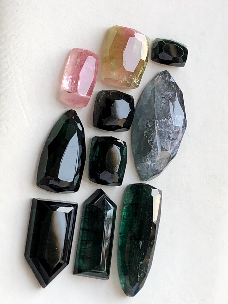 K&ouml;nnte beinhalten: Eine Sammlung von zehn facettierten Edelsteinen in verschiedenen Formen und Gr&ouml;&szlig;en. Die Steine zeigen Schattierungen von tiefem Schwarz, dunklem Waldgr&uuml;n, durchscheinendem Rosa und einem blassen Gelb-Rosa. Jeder Stein ist poliert.