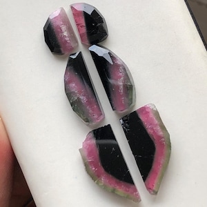 May include: Five pieces of watermelon tourmaline, a type of gemstone, in various shapes and sizes. The stones are black, pink, and green.