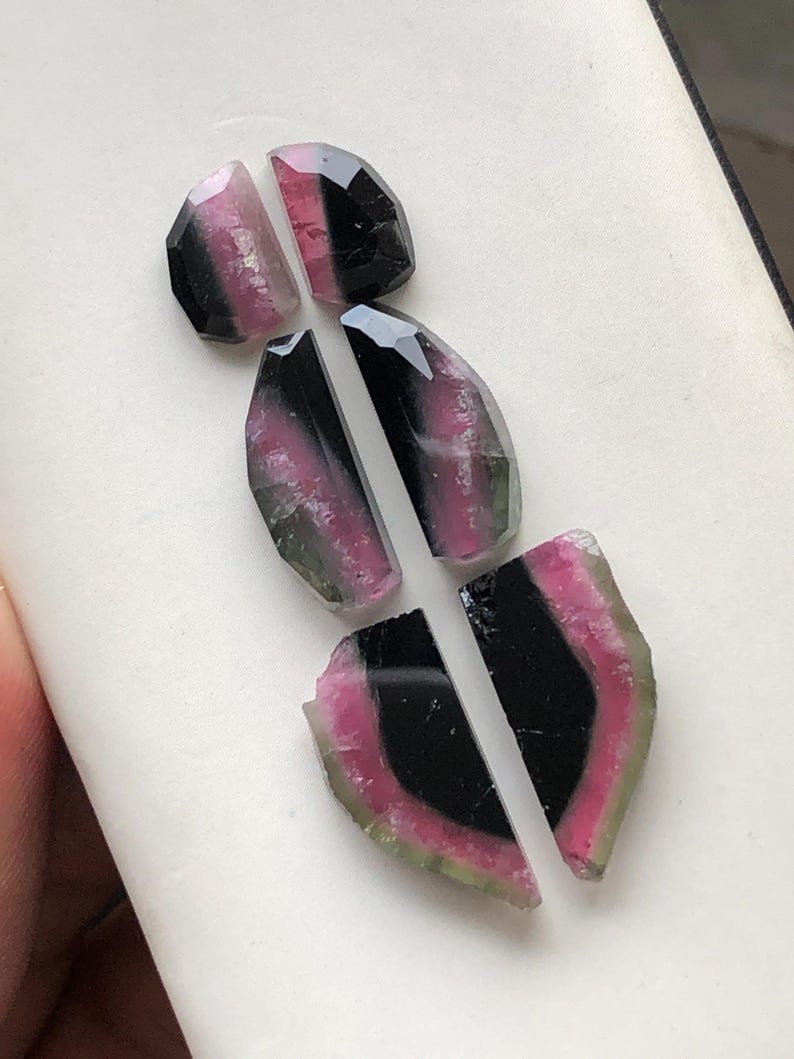 May include: Five pieces of watermelon tourmaline, a type of gemstone, in various shapes and sizes. The stones are black, pink, and green.