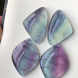 May include: Four polished fluorite gemstones, each in a rounded diamond shape. The stones show a blend of colours, including purple, green, and blue, with a glossy, reflective surface. The gemstones are arranged on a white surface.