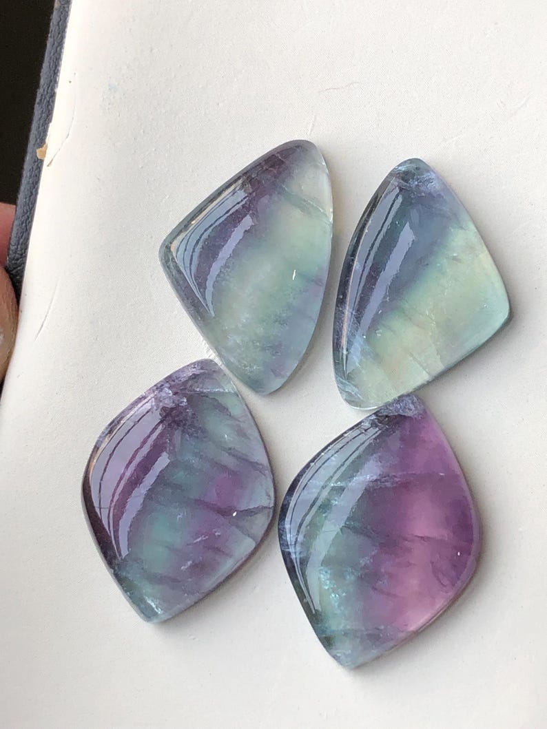 May include: Four polished fluorite gemstones, each in a rounded diamond shape. The stones show a blend of colours, including purple, green, and blue, with a glossy, reflective surface. The gemstones are arranged on a white surface.
