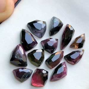 May include: A collection of 13 faceted, green, black, and pink tourmaline gemstones. The gemstones are cut in a teardrop shape and have a variety of sizes.