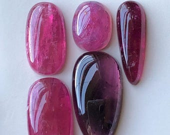 May include: Five polished gemstones in shades of pink and purple. The stones vary in shape, including oval and teardrop. The gems have a glossy, reflective surface, with visible internal textures and inclusions. The arrangement is on a white background.