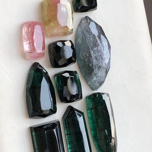 K&ouml;nnte beinhalten: Eine Sammlung von zehn facettierten Edelsteinen in verschiedenen Formen und Gr&ouml;&szlig;en, darunter Schattierungen in Rosa, Gelb, Schwarz und Dunkelgr&uuml;n. Die Steine haben polierte Oberfl&auml;chen mit geometrischen Schliffen und zeigen Farbvariationen.
