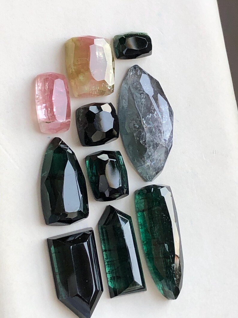 K&ouml;nnte beinhalten: Eine Sammlung von zehn facettierten Edelsteinen in verschiedenen Formen und Gr&ouml;&szlig;en, darunter Schattierungen in Rosa, Gelb, Schwarz und Dunkelgr&uuml;n. Die Steine haben polierte Oberfl&auml;chen mit geometrischen Schliffen und zeigen Farbvariationen.