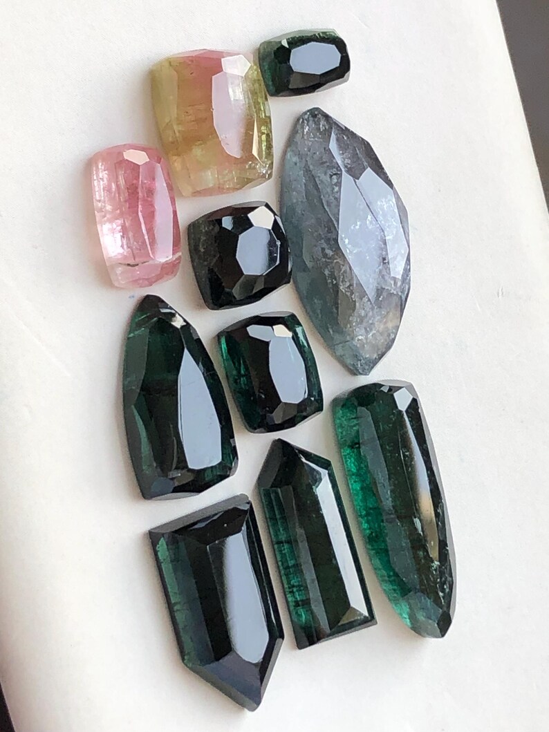 K&ouml;nnte beinhalten: Eine Sammlung von zehn facettierten Edelsteinen in verschiedenen Formen und Gr&ouml;&szlig;en. Die Steine zeigen eine Farbpalette von tiefem Waldgr&uuml;n, blassem Rosa, durchscheinendem Gelb bis hin zu dunklem Anthrazit, alle mit polierten Oberfl&auml;chen.