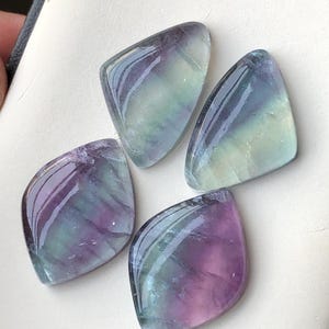 May include: Four polished fluorite gemstones in shades of purple, green, and blue. Each stone has a smooth, glossy surface and a unique shape, with rounded edges and a pointed top. The stones are arranged on a light-coloured surface.