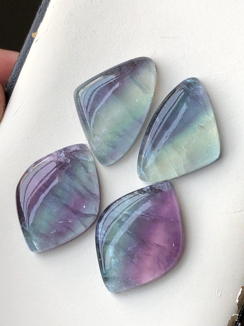 May include: Four polished fluorite gemstones in shades of purple, green, and blue. Each stone has a smooth, glossy surface and a unique shape, with rounded edges and a pointed top. The stones are arranged on a light-coloured surface.