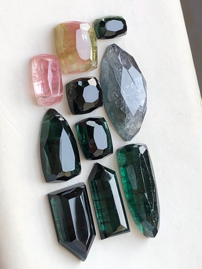 K&ouml;nnte beinhalten: Eine Sammlung von zehn facettierten Edelsteinen in verschiedenen Formen und Gr&ouml;&szlig;en. Die Steine zeigen eine Farbpalette aus tiefem Waldgr&uuml;n, durchscheinendem Rosa, blassem Gelb und dunklem Anthrazit mit polierten, geometrischen Schliffen.