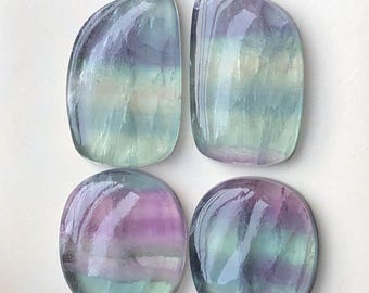 May include: Four polished fluorite gemstones in various shapes. Two are rounded rectangles, and two are oval. The stones display a mix of purple, green, and blue hues, with a glossy, translucent finish. The gemstones are set against a white background.