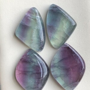 May include: Four polished fluorite gemstones, each with a diamond-like form. The stones exhibit a blend of purple, green, and blue hues, and have a glossy, reflective finish. The gemstones are presented on a light-coloured surface.
