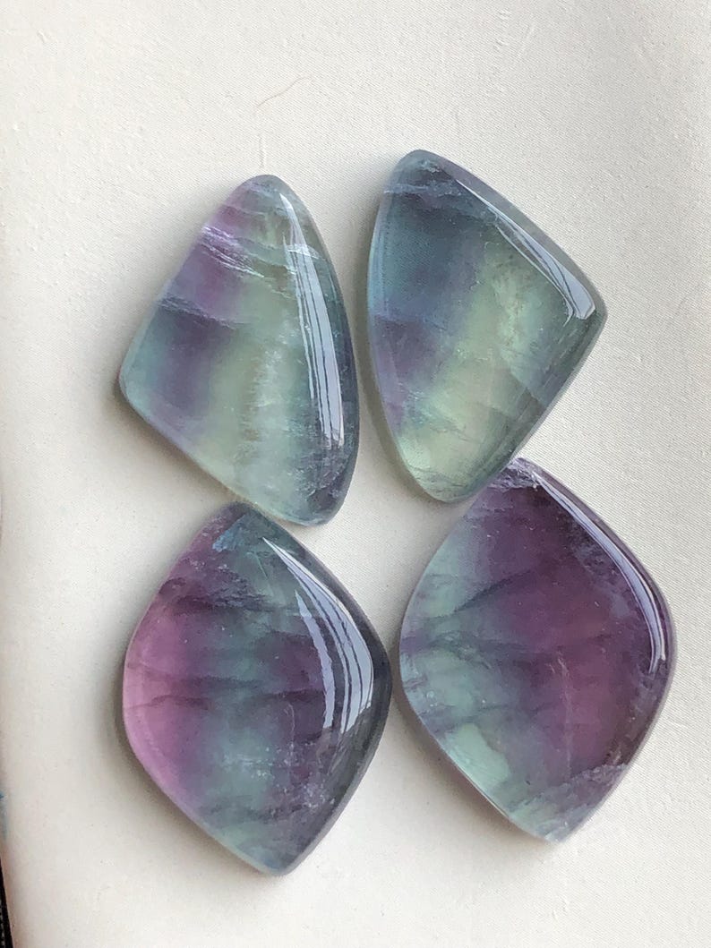 May include: Four polished fluorite gemstones, each with a diamond-like form. The stones exhibit a blend of purple, green, and blue hues, and have a glossy, reflective finish. The gemstones are presented on a light-coloured surface.