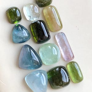 Puede incluir: Un surtido de piedras preciosas pulidas en varias formas y colores. Las gemas incluyen tonos de verde, azul y morado, con formas redondeadas y rectangulares. Las piedras tienen una superficie brillante y reflectante.