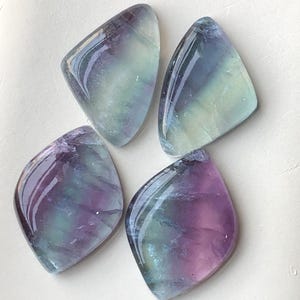 May include: Four polished fluorite gemstones in shades of purple, green, and blue. Each stone has a unique pattern and a glossy finish. The diamond-shaped stones, or cabochons, are arranged on a light background.