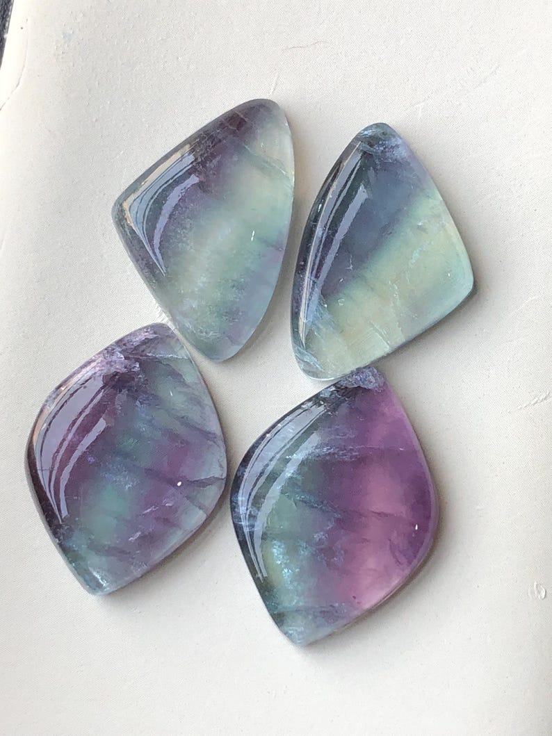 May include: Four polished fluorite gemstones in shades of purple, green, and blue. Each stone has a unique pattern and a glossy finish. The diamond-shaped stones, or cabochons, are arranged on a light background.