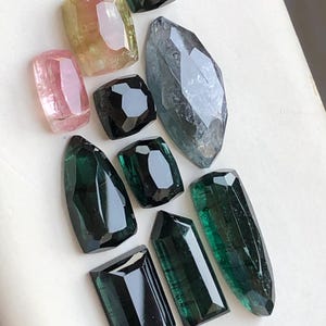 K&ouml;nnte beinhalten: Eine Sammlung von zehn facettierten Edelsteinen in verschiedenen Formen und Gr&ouml;&szlig;en. Das Sortiment umfasst rosa, gelbe und gr&uuml;ne Turmalinsteine sowie tiefschwarze und durchscheinend blaugraue Edelsteine, alle mit polierten Oberfl&auml;chen und Kanten.