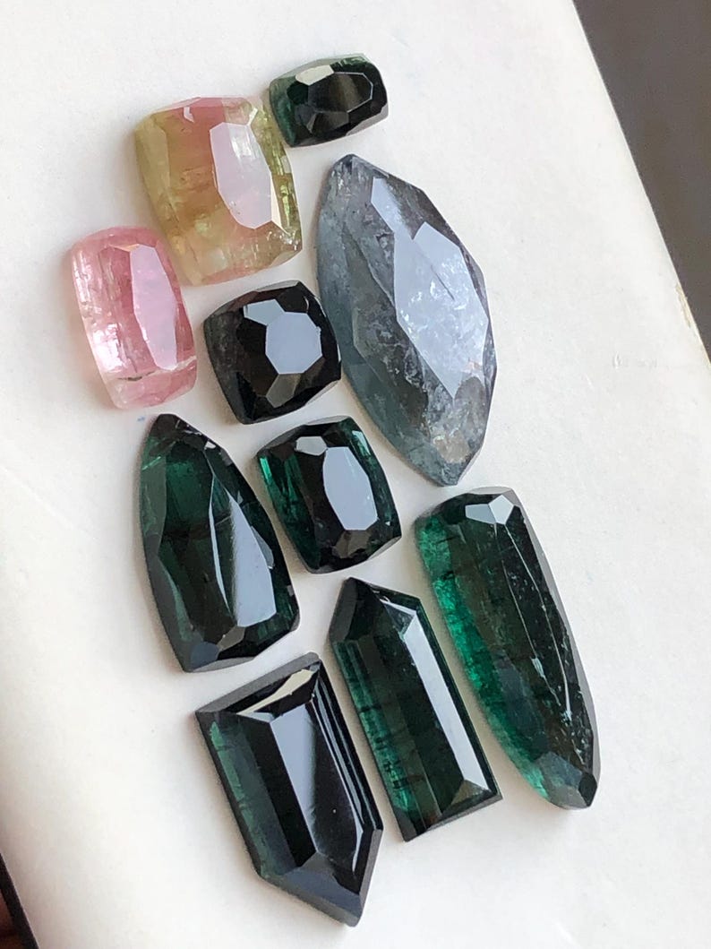 K&ouml;nnte beinhalten: Eine Sammlung von zehn facettierten Edelsteinen in verschiedenen Formen und Gr&ouml;&szlig;en. Das Sortiment umfasst rosa, gelbe und gr&uuml;ne Turmalinsteine sowie tiefschwarze und durchscheinend blaugraue Edelsteine, alle mit polierten Oberfl&auml;chen und Kanten.