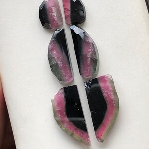 May include: Six pieces of watermelon tourmaline gemstone cabochons in various shades of pink, green, and black. The cabochons are cut in a variety of shapes, including ovals and half-ovals.