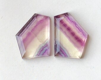 May include: Two faceted fluorite gemstones. Each stone has a unique pattern of purple, yellow, and clear bands. The gemstones are cut into a geometric shape with angled sides and a flat top.