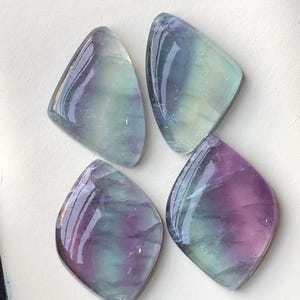 May include: Four polished fluorite gemstones. Each stone has a unique shape, resembling a rounded diamond or kite. The stones display a mix of colours, including purple, green, and clear, with a glossy finish. The gemstones are arranged on a white surface.
