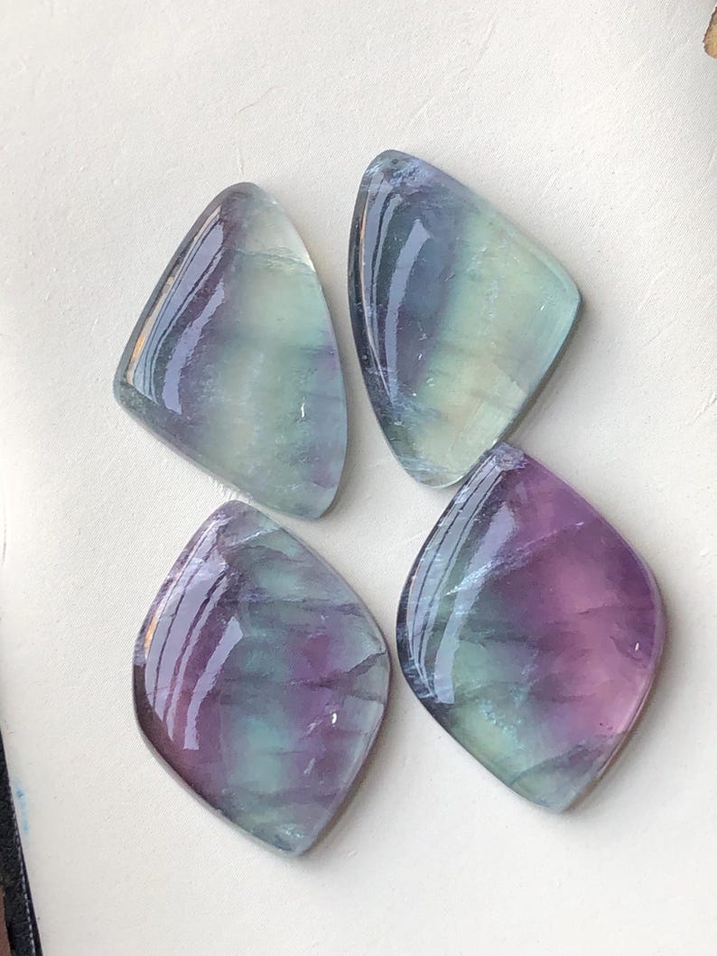May include: Four polished fluorite gemstones. Each stone has a unique shape, resembling a rounded diamond or kite. The stones display a mix of colours, including purple, green, and clear, with a glossy finish. The gemstones are arranged on a white surface.