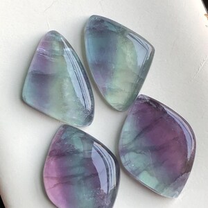 May include: Four polished fluorite gemstones in shades of purple, blue, and green. The stones have a smooth, glossy surface and are irregularly shaped. The gemstones are arranged on a white surface.