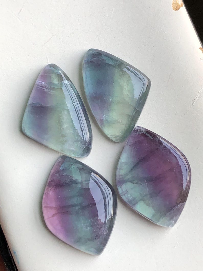 May include: Four polished fluorite gemstones in shades of purple, blue, and green. The stones have a smooth, glossy surface and are irregularly shaped. The gemstones are arranged on a white surface.