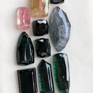 K&ouml;nnte beinhalten: Eine Sammlung von zehn facettierten Edelsteinen in verschiedenen Formen und Gr&ouml;&szlig;en. Die Steine zeigen Schattierungen in Rosa, Gelb, Dunkelgr&uuml;n, Schwarz und einen gro&szlig;en, durchscheinenden grauen Stein im Marquise-Schliff auf wei&szlig;em Grund.