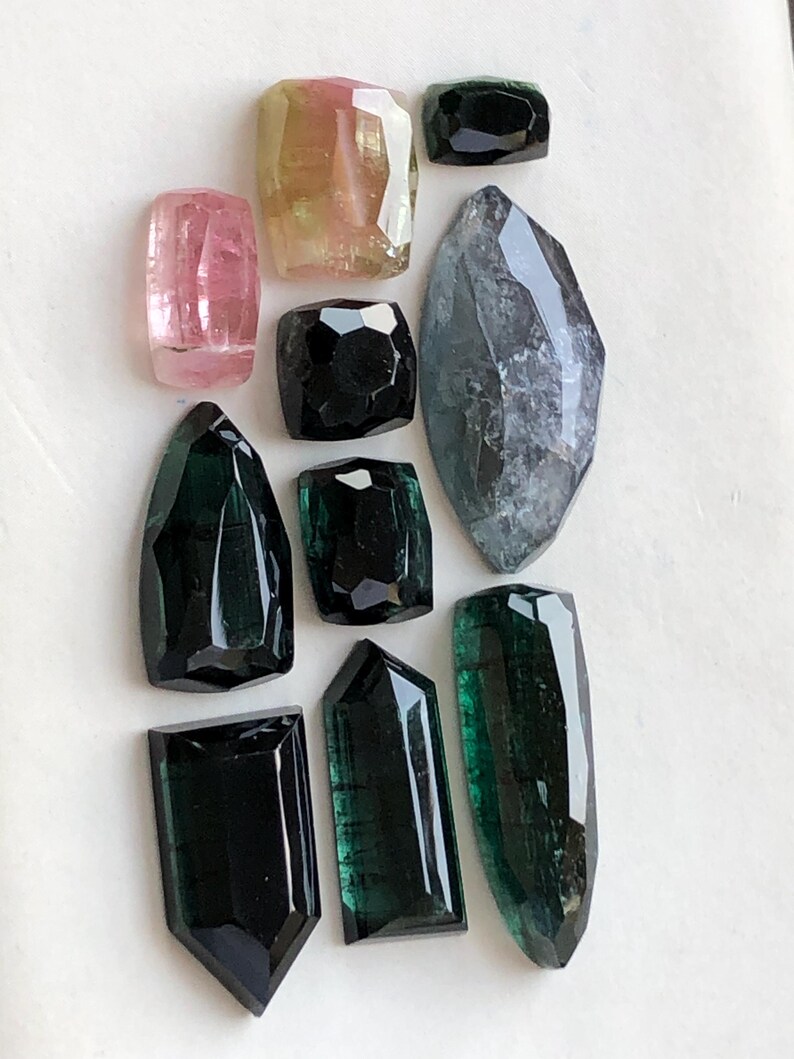 K&ouml;nnte beinhalten: Eine Sammlung von zehn facettierten Edelsteinen in verschiedenen Formen und Gr&ouml;&szlig;en. Die Steine zeigen Schattierungen in Rosa, Gelb, Dunkelgr&uuml;n, Schwarz und einen gro&szlig;en, durchscheinenden grauen Stein im Marquise-Schliff auf wei&szlig;em Grund.