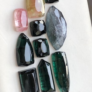 K&ouml;nnte beinhalten: Eine Sammlung von zehn facettierten Edelsteinen in verschiedenen Formen und Gr&ouml;&szlig;en, darunter T&ouml;ne in Rosa, Gelb, Schwarz, Dunkelgr&uuml;n und ein gro&szlig;er, durchscheinender grauer Stein im Markisenschliff, angeordnet auf einer wei&szlig;en Fl&auml;che.