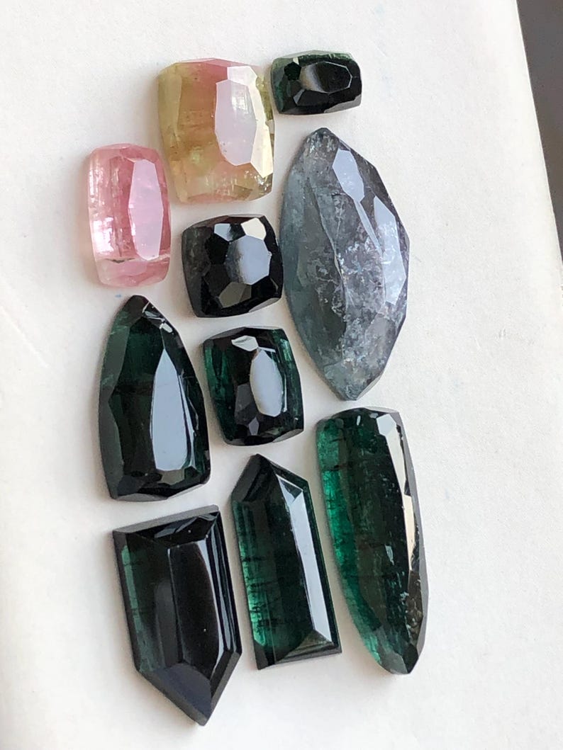 K&ouml;nnte beinhalten: Eine Sammlung von zehn facettierten Edelsteinen in verschiedenen Formen und Gr&ouml;&szlig;en, darunter T&ouml;ne in Rosa, Gelb, Schwarz, Dunkelgr&uuml;n und ein gro&szlig;er, durchscheinender grauer Stein im Markisenschliff, angeordnet auf einer wei&szlig;en Fl&auml;che.
