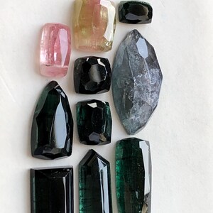 K&ouml;nnte beinhalten: Eine Sammlung von zehn facettierten Edelsteinen in verschiedenen Formen und Gr&ouml;&szlig;en, darunter T&ouml;ne in Rosa, Gelb, Schwarz, Dunkelgr&uuml;n und ein gro&szlig;er, durchscheinender grauer Stein im Marquise-Schliff, angeordnet auf einer wei&szlig;en Fl&auml;che.