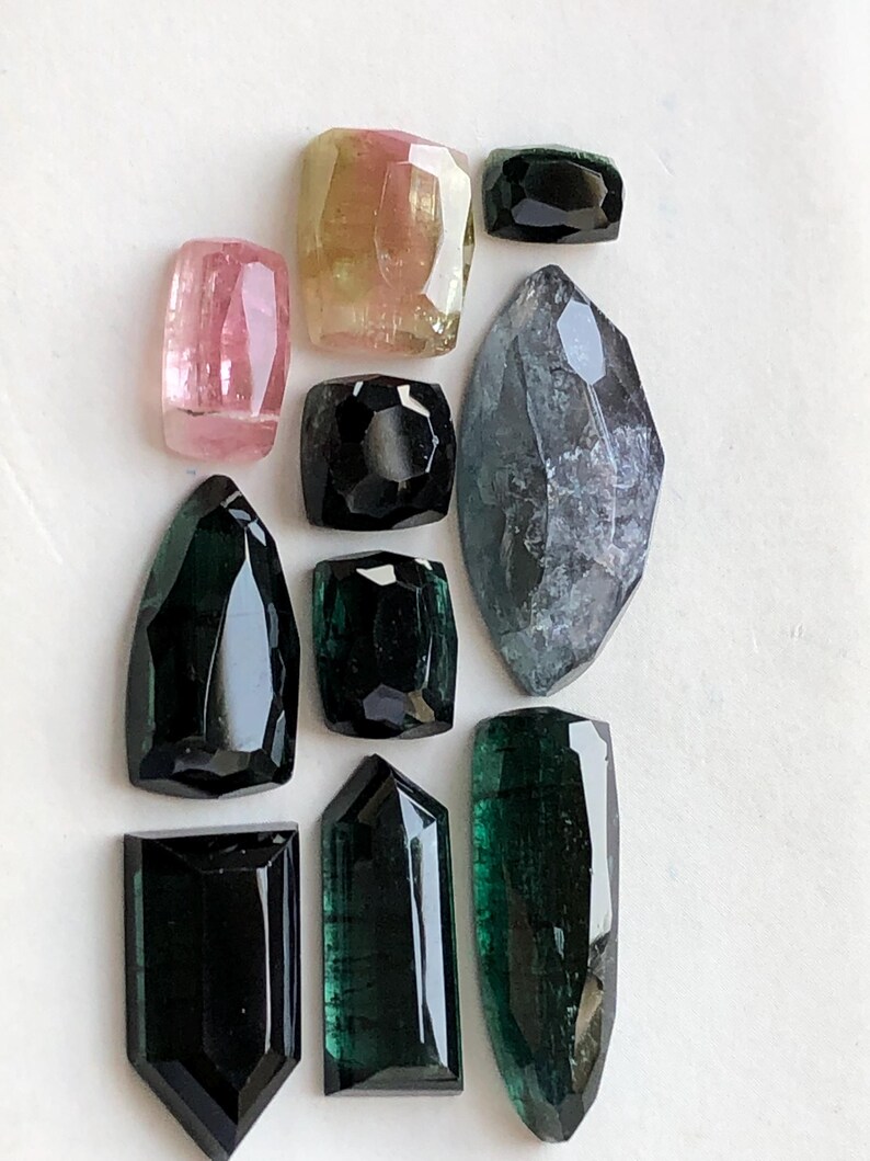 K&ouml;nnte beinhalten: Eine Sammlung von zehn facettierten Edelsteinen in verschiedenen Formen und Gr&ouml;&szlig;en, darunter T&ouml;ne in Rosa, Gelb, Schwarz, Dunkelgr&uuml;n und ein gro&szlig;er, durchscheinender grauer Stein im Marquise-Schliff, angeordnet auf einer wei&szlig;en Fl&auml;che.